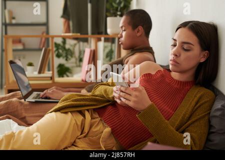 Brünette junge Frau beleidigt von Freundin auf dem Bett liegen und Surfen Websites auf dem Smartphone Stockfoto