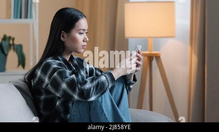 Seitenansicht der traurig gelangweilten asiatischen Frau Mädchen sitzt auf dem Sofa zu Hause mit Telefon serious unglücklich Dame Scrollen im Smartphone und mit Handy Stockfoto