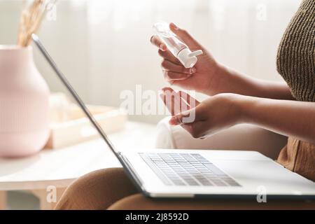 Nahaufnahme einer nicht erkennbaren Frau, die mit einem Laptop auf den Knien sitzt und Antiseptikum in die Hand gießt, während sie es während des Coronavirus reinigt Stockfoto