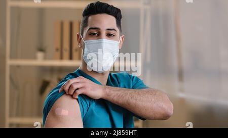 Junger arabischer Mann Praktizierender Arzt Krankenschwester stehen innen in schützender chirurgischer Gesichtsmaske zeigen klebende Verband auf der Schulter nach dem Impfstoff Stockfoto