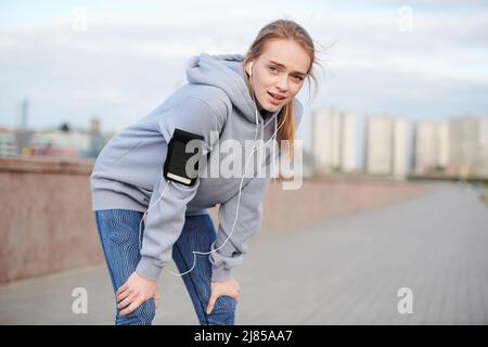 Porträt einer sportlichen jungen Frau mit einem Armband, das sich auf die Knie stützt, während sie sich nach einem erschöpfenden Lauf über die Stadt ausruhte Stockfoto