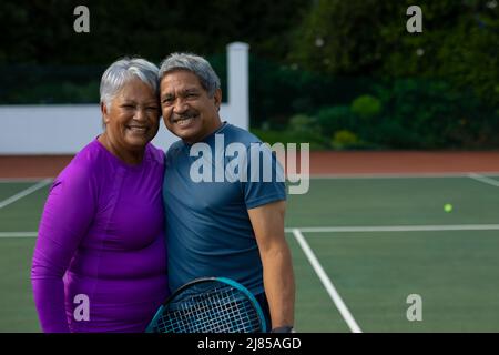 Porträt eines lächelnden Senioren-Paares, das sich auf dem Tennisplatz umarmt Stockfoto