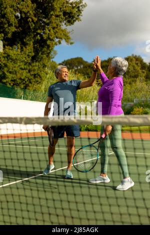 Glückliches Senioren-Paar mit Biracial, das Schläger hält und auf dem Tennisplatz gegen den bewölkten Himmel High-Five-Schläger gibt Stockfoto