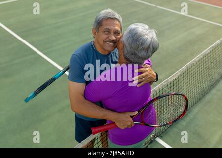 Hochwinkelporträt eines älteren Birazialmannes, der eine Frau umarmt, während er am Tennisnetz auf dem Tennisplatz steht Stockfoto