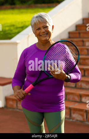 Porträt einer lächelnden älteren Frau aus der Birazialzeit mit kurzen Haaren, die Tennisschläger gegen die Stufen hält Stockfoto