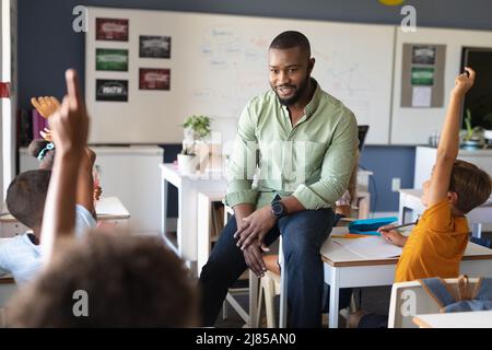 afroamerikanischer junger männlicher Lehrer, der multirassische Studenten ansieht, die während des Unterrichts die Hände heben Stockfoto