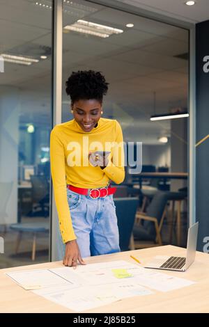 Lächelnde afroamerikanische Geschäftsfrau, die auf einem Smartphone-Lautsprecher spricht, während sie nach Dokumenten schaut Stockfoto
