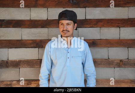 Ein gut aussehender indischer junger Mann, der eine Kappe nach hinten trägt und seitlich blickt, während er gegen eine Holz- und Steinwand steht Stockfoto
