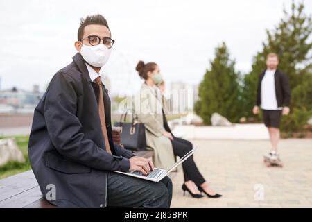 Porträt eines hübschen arabischen Mannes mit Brille und Maske, der im Stadtpark auf der Bank sitzt und auf dem Laptop tippt Stockfoto