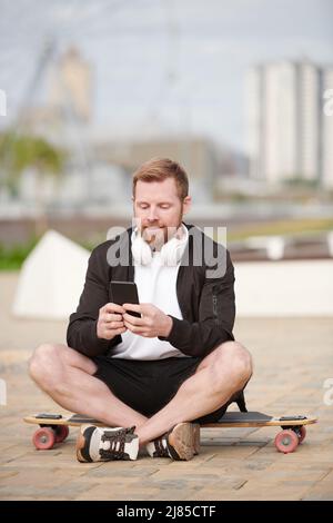 Content junge bärtige Mann in Jacke sitzt auf dem Skateboard und SMS-Nachricht auf Telefon im Freien Stockfoto