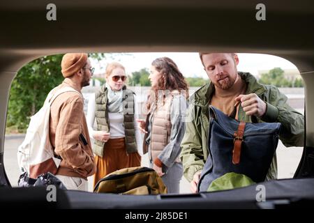 Bärtiger Mann in einer Jacke, der Sachen in den Kofferraum packte, während seine Freunde über Reisen mit dem Auto sprachen Stockfoto