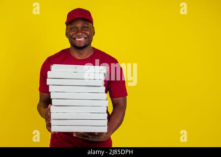 afroamerikanischer Lieferer mit Paket, das rotes T-Shirt und Mütze auf gelbem Hintergrund trägt. Konzept für Fast Delivery Food Stockfoto