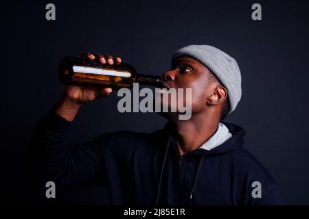 afroamerikanische gue trägt grauen Hut und Hoody hjlding Botle mit Wein in srudio schwarzen Hintergrund.Alkoholismus Obdachlosen bum Konzept Stockfoto