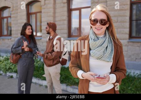 Porträt einer lächelnden jungen Frau in einer Sonnenbrille mit Smartphone-App im Freien, während ihre Freunde im Hintergrund chatten Stockfoto