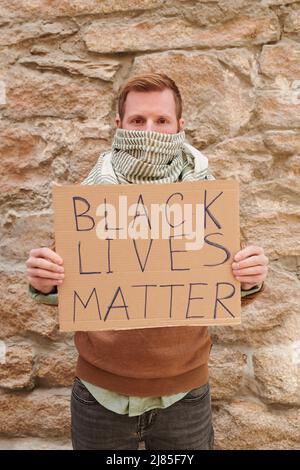 Porträt eines jungen Mannes mit verdeckter Nase und Mund und Schal, der das Banner „Black Lives Matter“ zeigt, während er an einem Protest teilnimmt Stockfoto