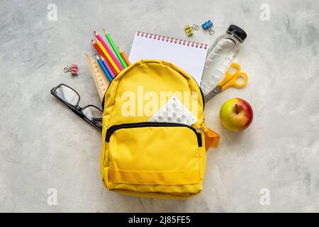 Zurück zum Schulkonzept. Gelber Schulrucksack mit Vorräten Wasser und Apfel Stockfoto