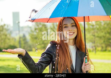 Rothaarige Ingwerfrau, die im Sommerpark unter einem bunten Regenschirm steht und nach Regenbogen prüft Stockfoto