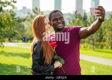 Nahaufnahme eines interracial Paares mit weißer Rose im Herbstpark im Frühjahr, das ein Selfie mit einer Smartphone-Kamera gemacht hat Stockfoto