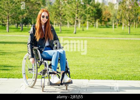 Rothaarige Ingwerfrau, die glücklich ist, sie sitzt im Rollstuhl, trägt eine Sonnenbrille und eine warme Lederjacke im Sommerpark Stockfoto