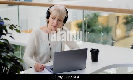 Reife kaukasische Frau Geschäftsfrau Lehrer in Kopfhörern schaut auf Laptop-Bildschirm nimmt Notizen schreibt Bericht Abstand lernen online Nachhilfe studieren Stockfoto