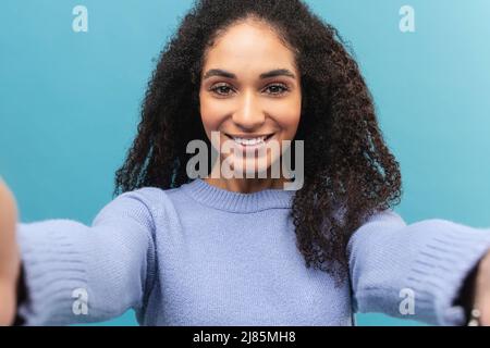 Sorglose afroamerikanische junge Frau mit lockigen Haaren, die Selfie auf dem Smartphone macht und Inhalte für soziale Medien macht, isoliert auf Blau Stockfoto