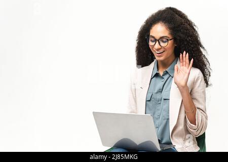 Selbstbewusste multirassische Frau in Smart Wear mit Laptop isoliert auf weißem Hintergrund. Weibliche Angestellte oder Studentin winkt zum Bildschirm und hat einen Videoanruf Stockfoto
