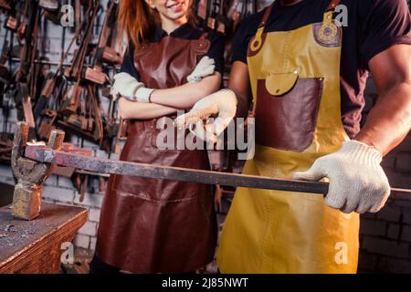 Verliebtes Paar rothaarige junge europäische Frau und afroamerikanischer Mann mit Lederschürze, die Schmiede in der Werkstatt arbeitet.kleine Familie Stockfoto