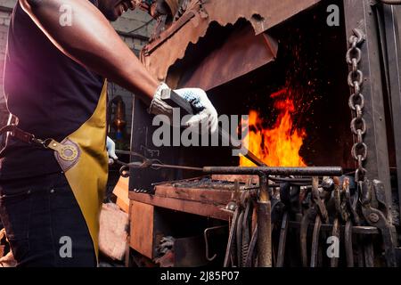 Schöner afrikanischer amerikanischer Mann schmiedet Stahl neben dem Ofen in dunkler Werkstatt. Kleinbetrieb Stockfoto