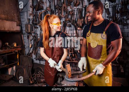 Verliebtes Paar rothaarige junge europäische Frau und afroamerikanischer Mann mit Lederschürze, die Schmiede in der Werkstatt arbeitet.kleine Familie Stockfoto