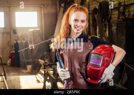 Stark und würdig ziemlich Rotschopf Ginher Frau trägt Schutzhelm und Lederschürze mit Handschuhen hält Schweißmaschine Instrument . Klein Stockfoto