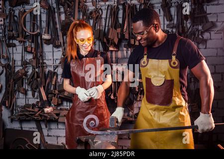 Verliebtes Paar rothaarige junge europäische Frau und afroamerikanischer Mann mit Lederschürze, die Schmiede in der Werkstatt arbeitet.kleine Familie Stockfoto