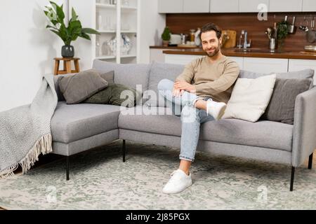 Porträt eines jungen kaukasischen Mannes, der auf dem Sofa ruht. Schöner positiver Mann auf dem Sofa sitzt im Wohnzimmer, träumt und entspannt zu Hause Stockfoto