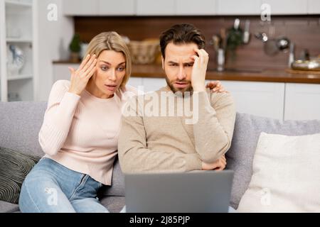 Junge Ehepartner, die die Hypothek nicht bezahlen können. Frustrierte Ehefrau und verärgerte Ehemann, die Rechnungen auf dem Laptop durchschauen. Junges kaukasisches Paar mit finanziellen Problemen Stockfoto