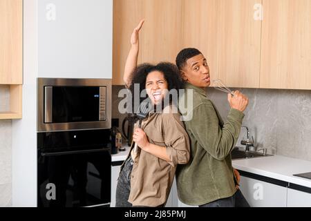 Junge multirassische Frau und hübscher Mann lächeln und haben Spaß zusammen, halten Küchenutensilien. Glückliches interrassisches Paar, das in der Küche tanzt und beim Kochen des Frühstücks oder Abendessens singt Stockfoto