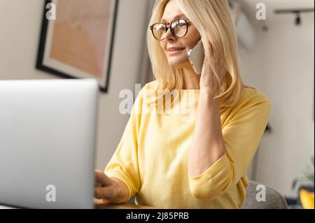 Lächelnd freundliche ältere Frau arbeitet von zu Hause aus. Sie spricht am Telefon, während sie am Tisch vor dem Laptop und Notebook sitzt Stockfoto