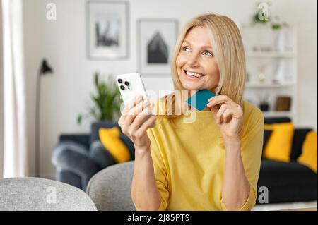 Glückliche pensionierte Frau gibt Ersparnisse für Online-Shopping aus. Fröhliche ältere Frau hält Smartphone und Kreditkarte in den Händen und macht Kauf online, mit mobilen App für den Kauf Stockfoto
