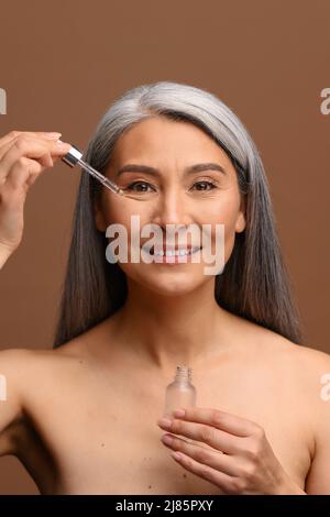 Vertikales Porträt einer lächelnden asiatischen Frau mittleren Alters mit langen grauen Haaren, die ein Glas mit Serum in den Händen hält und Pipette zum Auftragen des Kosmetikprodukts verwendet Stockfoto