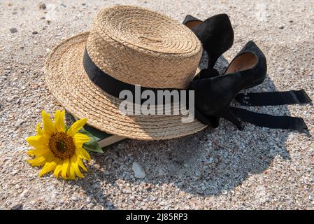 Nahaufnahme von gelbem Strohhut, Sonnenblume, schwarzen Schuhen mit hohen Absätzen für Frauen und grünem Buch, das auf dem Meersand liegt, Blick aus dem hohen Winkel. Relax and Rest Konzept. Stockfoto