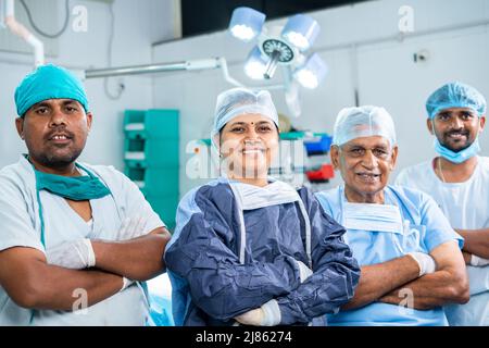 Gruppe von lächelnden erfolgreichen Chirurgen, die selbstbewusst stehen, indem sie die Kamera im Operationssaal betrachten - ein Gesteiner von Spezialisten im Gesundheitswesen, Fachwissen und Stockfoto