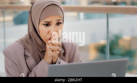 Traurige muslimische Studentin in Hijab, die E-Mails auf dem Laptop-Bildschirm liest, schockiert durch schlechte Nachrichten frustrierte Geschäftsfrau sorgt sich um finanzielle Probleme Stockfoto