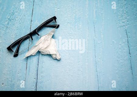 Draufsicht auf Lesebrille mit schwarzen Bügeln und zerknittertem Brillenreinigungstuch auf hellblau lackierter Holzoberfläche Stockfoto