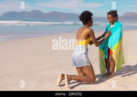 afroamerikanische junge Mutter wickelte glückliche Tochter mit einem Handtuch, während sie am Strand gegen den Himmel kniete Stockfoto
