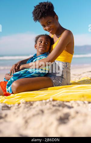 Porträt einer lächelnden afroamerikanischen jungen Frau, die mit ihrer Tochter in einem Handtuch auf einer Decke sitzt Stockfoto