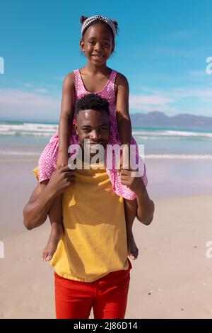 Porträt eines lächelnden afroamerikanischen jungen Mannes, der seine Tochter auf den Schultern gegen Meer und Himmel trägt Stockfoto