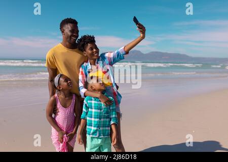 Glückliche afroamerikanische junge Frau, die am Strand Selfie mit Mann, Sohn und Tochter über das Smartphone macht Stockfoto