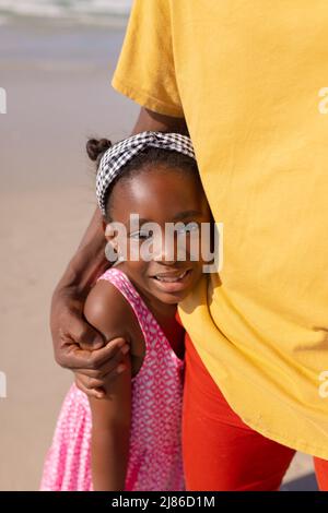 Mittelteil eines afroamerikanischen jungen Mannes, der seine lächelnde Tochter umarmt, während er am Strand steht Stockfoto