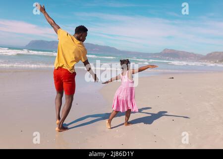 Rückansicht des verspielten afroamerikanischen jungen Mannes und seiner Tochter mit ausgestreckten Armen am Strand Stockfoto