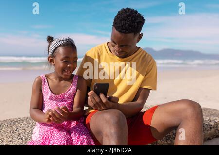 afroamerikanischer junger Mann mit Mobiltelefon, während er mit seiner Tochter am Strand gegen den Himmel sitzt Stockfoto