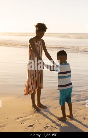 afroamerikanische junge Frau, die die Hände des Sohnes hält, während sie bei Sonnenuntergang am Strand gegen den klaren Himmel steht Stockfoto