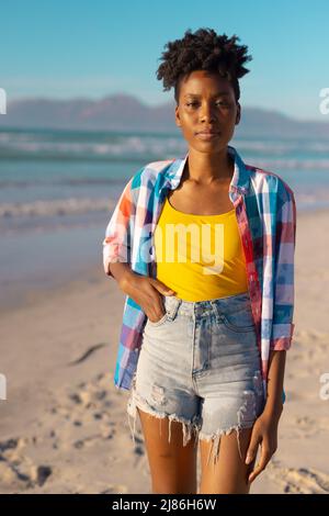 Porträt einer afroamerikanischen jungen Frau mit kurzen Haaren und Hand in der Tasche gegen Meer und blauen Himmel Stockfoto